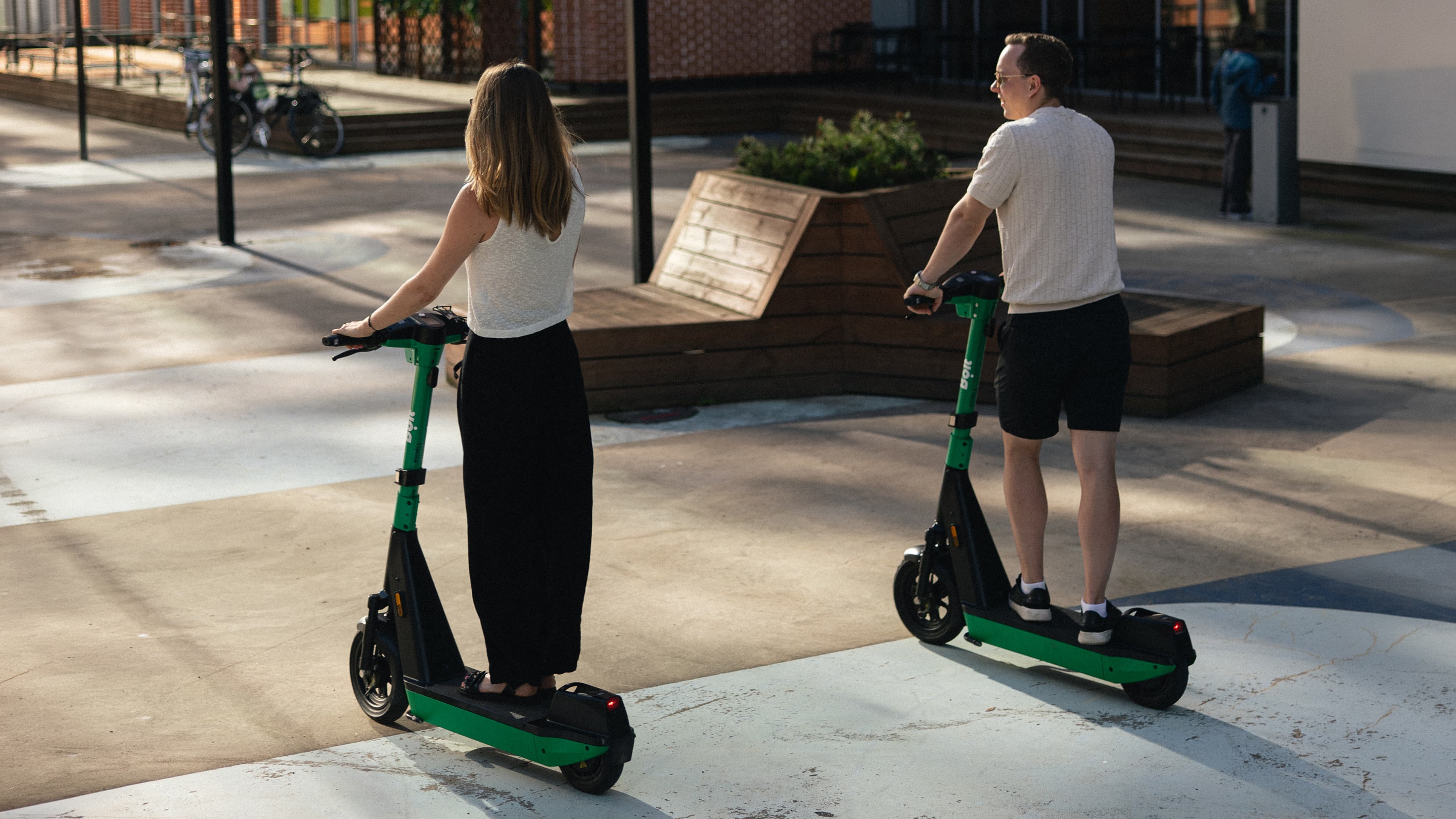 Two people riding Bolt scooters across a paved square