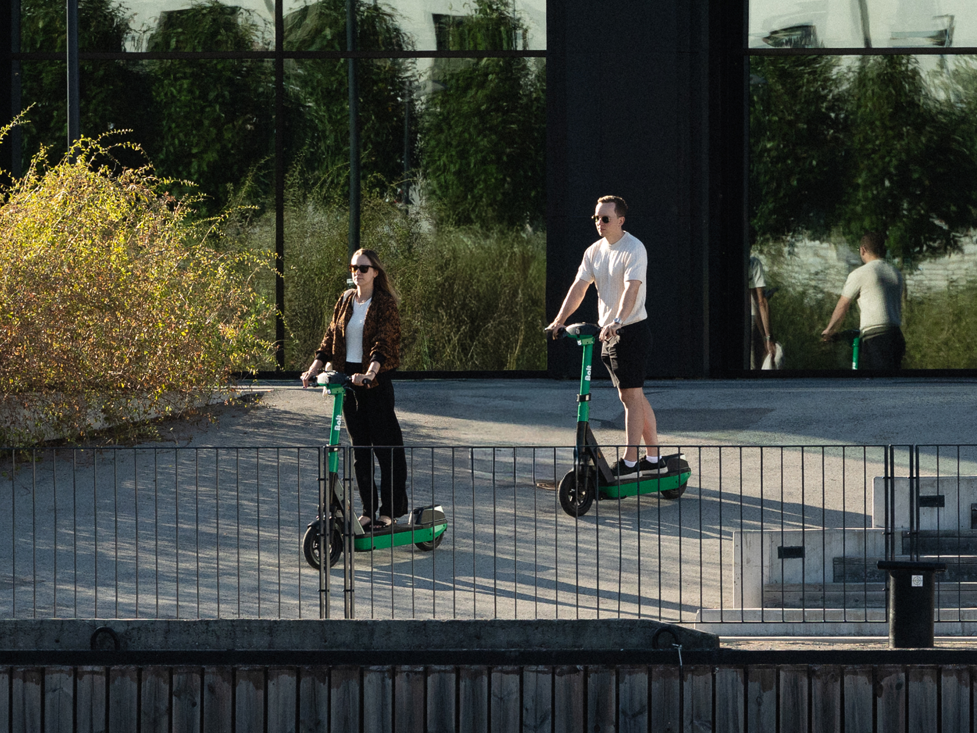 Two Bolt scooter riders on a terrace beside a glass building